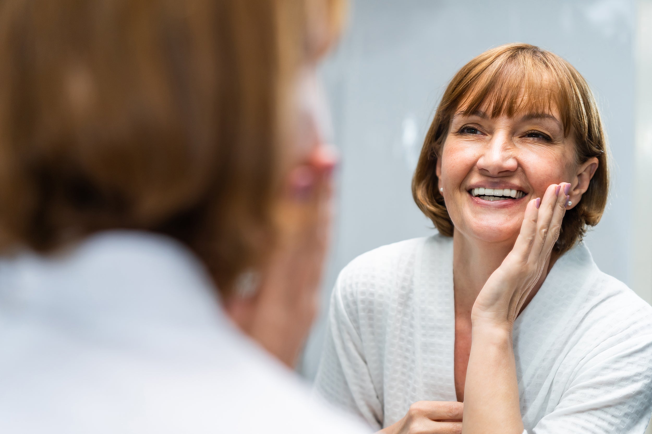 An older woman looks at her face in the mirror. Wondering how long it takes for scars to fade after cosmetic surgery? Get the full 12–18 month healing timeline, stage by stage, plus what affects it.