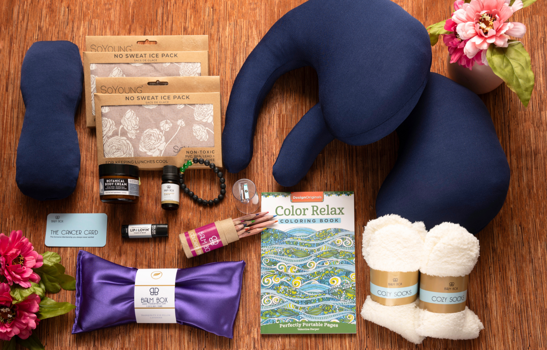 A flat lay of self-care items on a wood surface, including a U-shaped neck pillow, coloring book, ice packs, soft socks, eye mask, lotion, bracelet, flowers, and colored pencils.