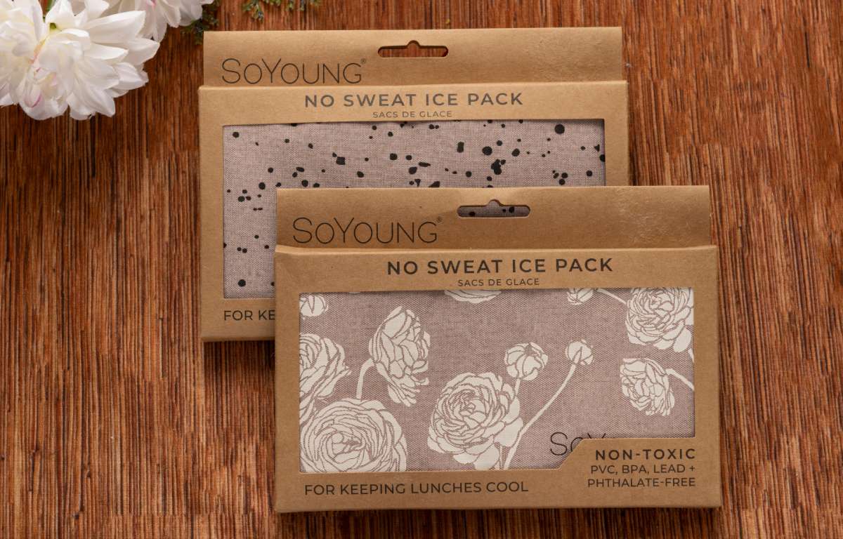 Two rectangular SoYoung No Sweat Ice Packs—one with a black splatter design, another with a white floral pattern—are displayed on wood with white flowers nearby, included in The Best Mastectomy Care Package.
