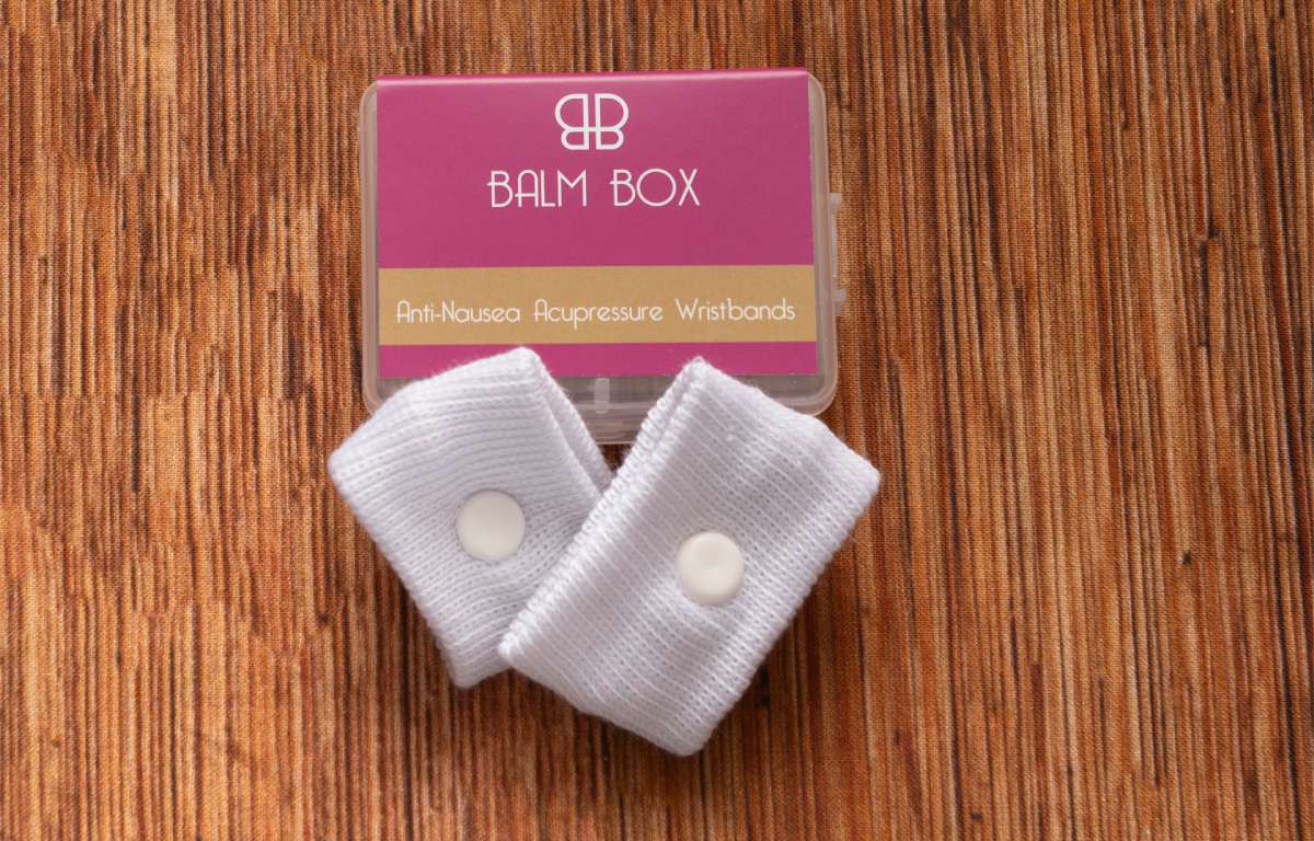 A pair of white acupressure wristbands for nausea relief sit before a pink and gold Balm Box case labeled “Anti-Nausea Acupressure Wristbands," as part of The Perfect Little Anti-Nausea Box—a thoughtful cancer patient gift on a wooden surface.
