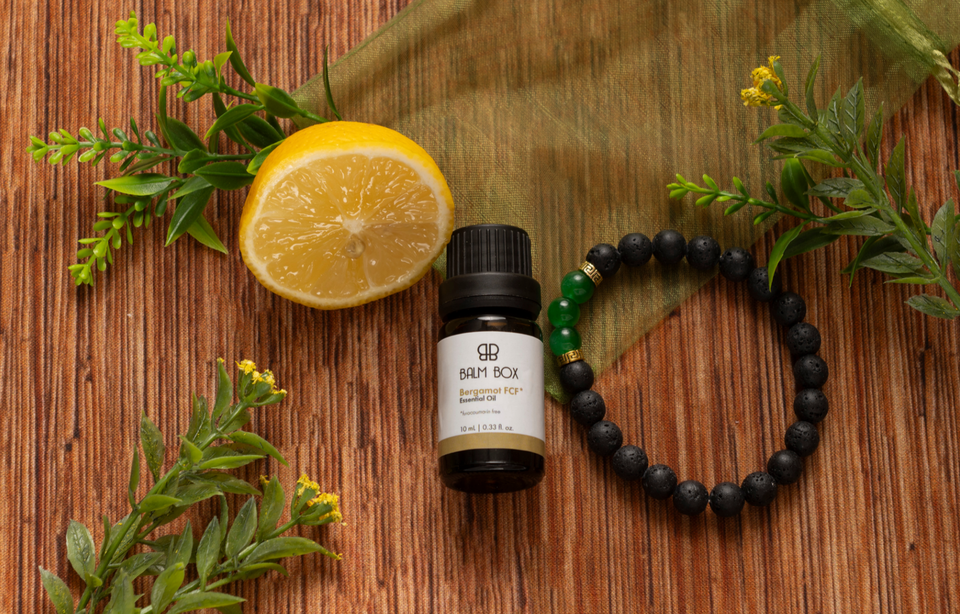 A bottle of essential oil from The Best Mastectomy Care Package rests on wood, surrounded by a lemon half, black bead bracelet, green cloth, and leafy sprigs—adding an uplifting touch for self-care or mastectomy recovery.