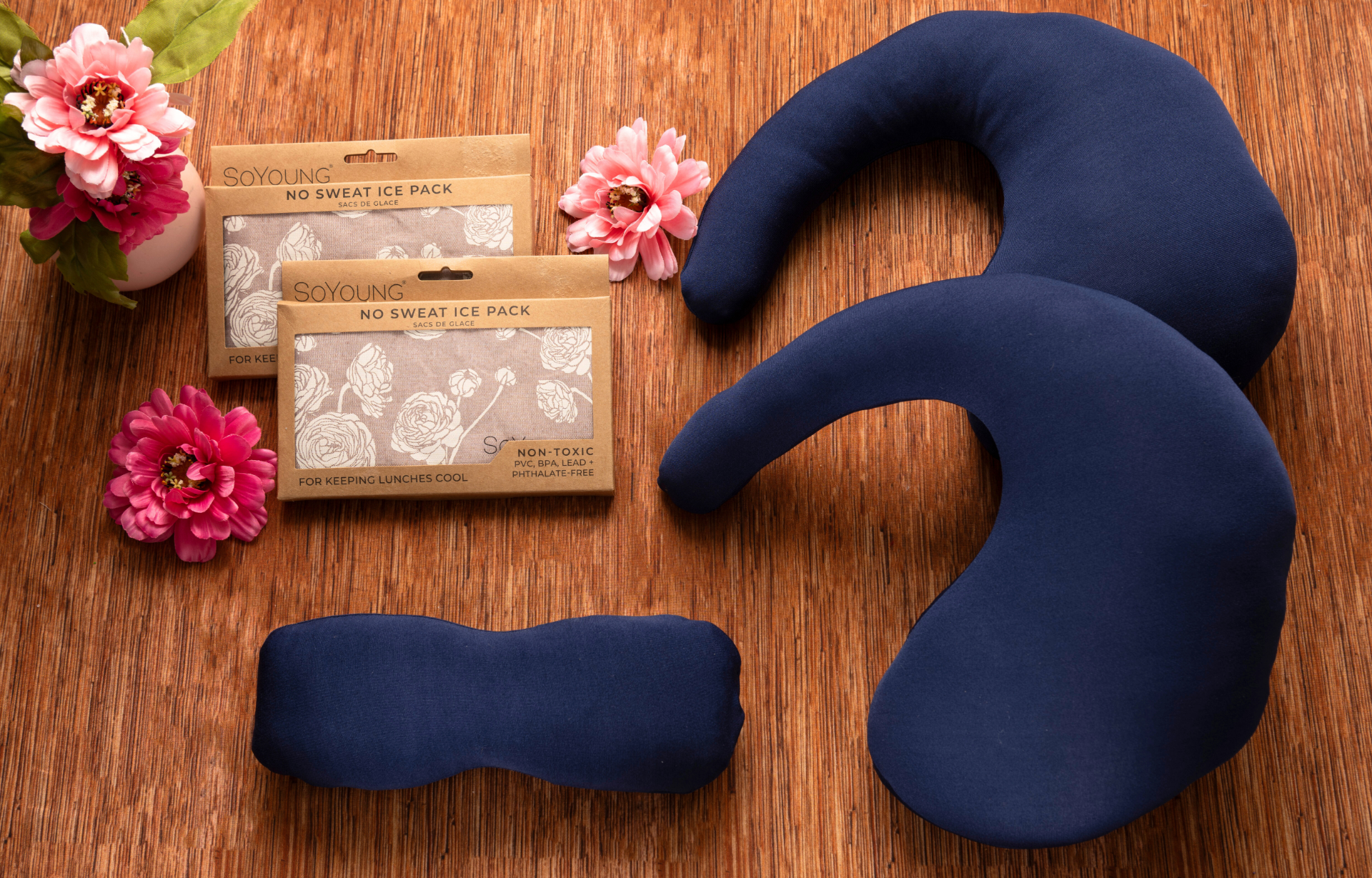 The Best Mastectomy Care Package features navy blue therapeutic pillows, a small cushion, and two floral ice pack boxes on a wooden surface with pink artificial flowers in a vase—ideal for self-care or gifting to cancer patients.