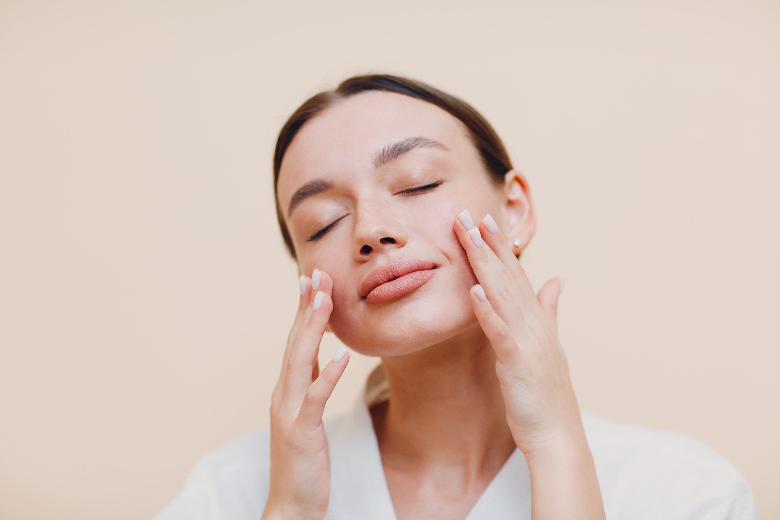 A woman with closed eyes gently touching her cheeks with both hands, appearing relaxed and content, against a neutral beige background. She has neatly groomed eyebrows and natural-looking makeup.