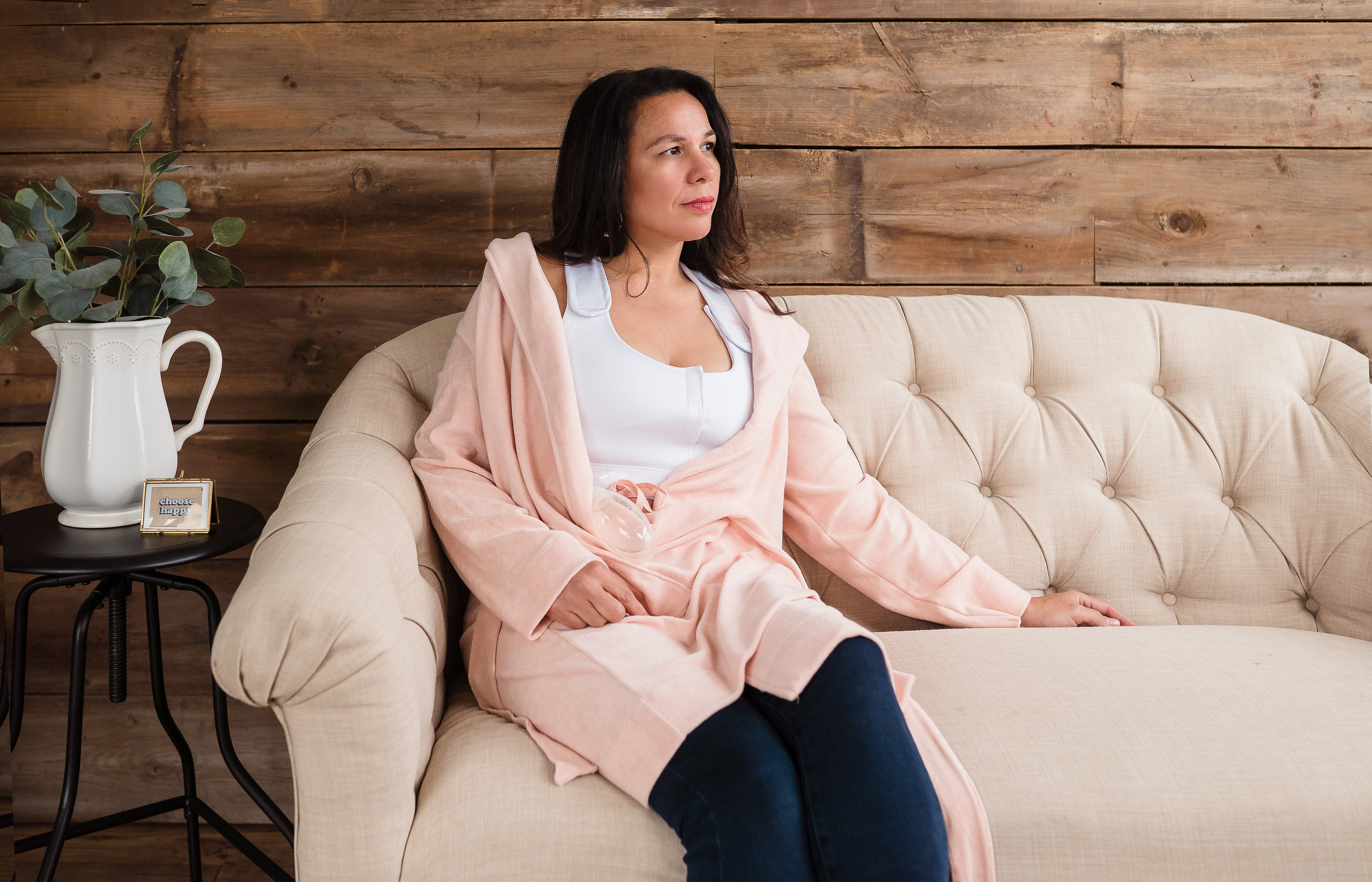 A woman in a light pink robe and dark pants sits on a cream-colored tufted sofa, suggesting the comfort of the Larissa Post-Surgical Bra for breast surgery recovery, with greenery nearby adding to the peaceful setting.
