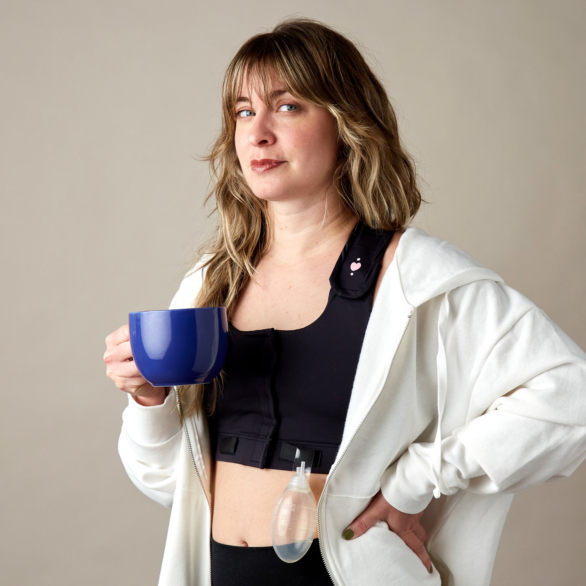 A woman with long hair wears a white hoodie over a black Larissa Post-Surgical Bra, holding a large blue mug. A surgical drain bulb is attached to her vest, with a medical dressing on her chest. She looks confidently at the camera.