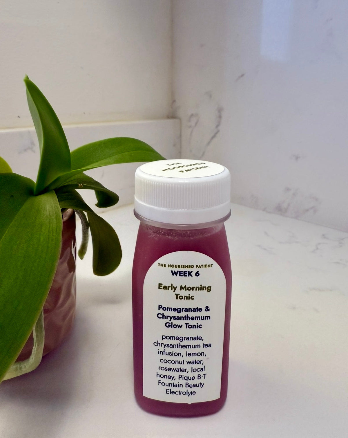 A small plastic bottle labeled Early Morning Tonic, containing pink liquid, sits on a white countertop near a green plant. The label highlights pomegranate and chrysanthemum tea, part of the 7-Day Facelift Recovery Meal Program to help reduce swelling.