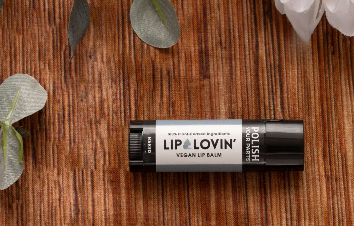 A black tube of Lip Lovin vegan lip balm rests on a brown striped surface with green leaves and a white flower—featuring plant-derived ingredients, it's a soothing addition to The Best Mastectomy Care Package or any self-care box.