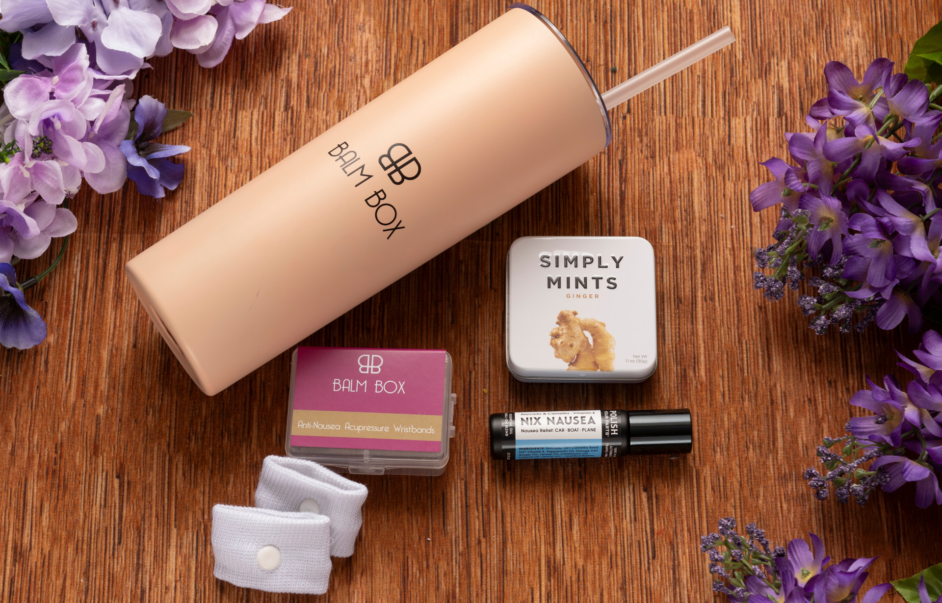 The Perfect Little Anti-Nausea Box features a peach tumbler, Simply Mints tin, No Mo Nausea roll-on, two acupressure wristbands in a pink BAM BOX case—thoughtfully arranged to ease nausea and chemotherapy side effects—plus purple flowers.