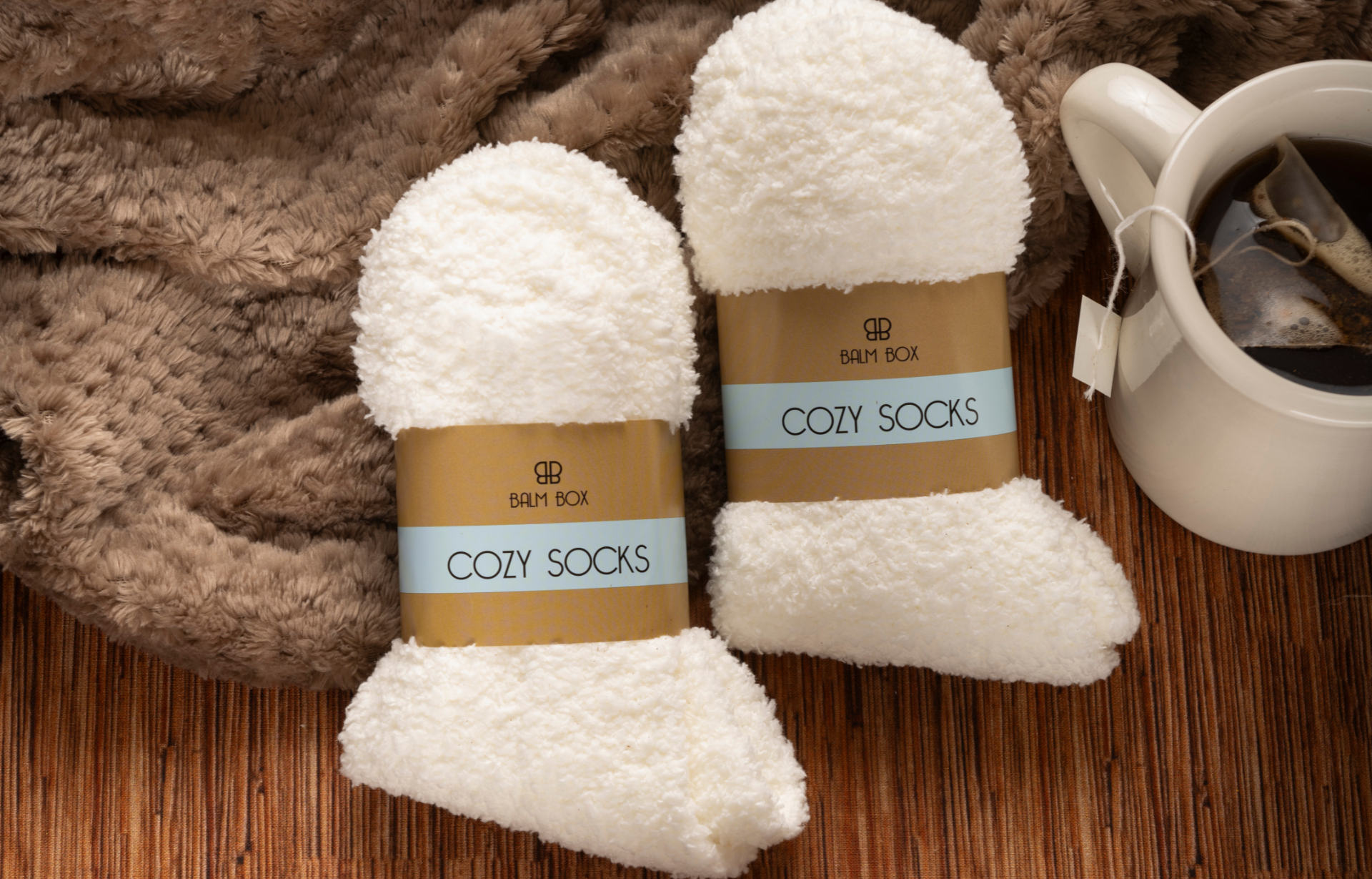 Two pairs of fluffy white socks from The Best Mastectomy Care Package rest on a brown textured surface next to a fuzzy blanket and tea, making them an ideal choice for any self-care box or cancer patient gift.