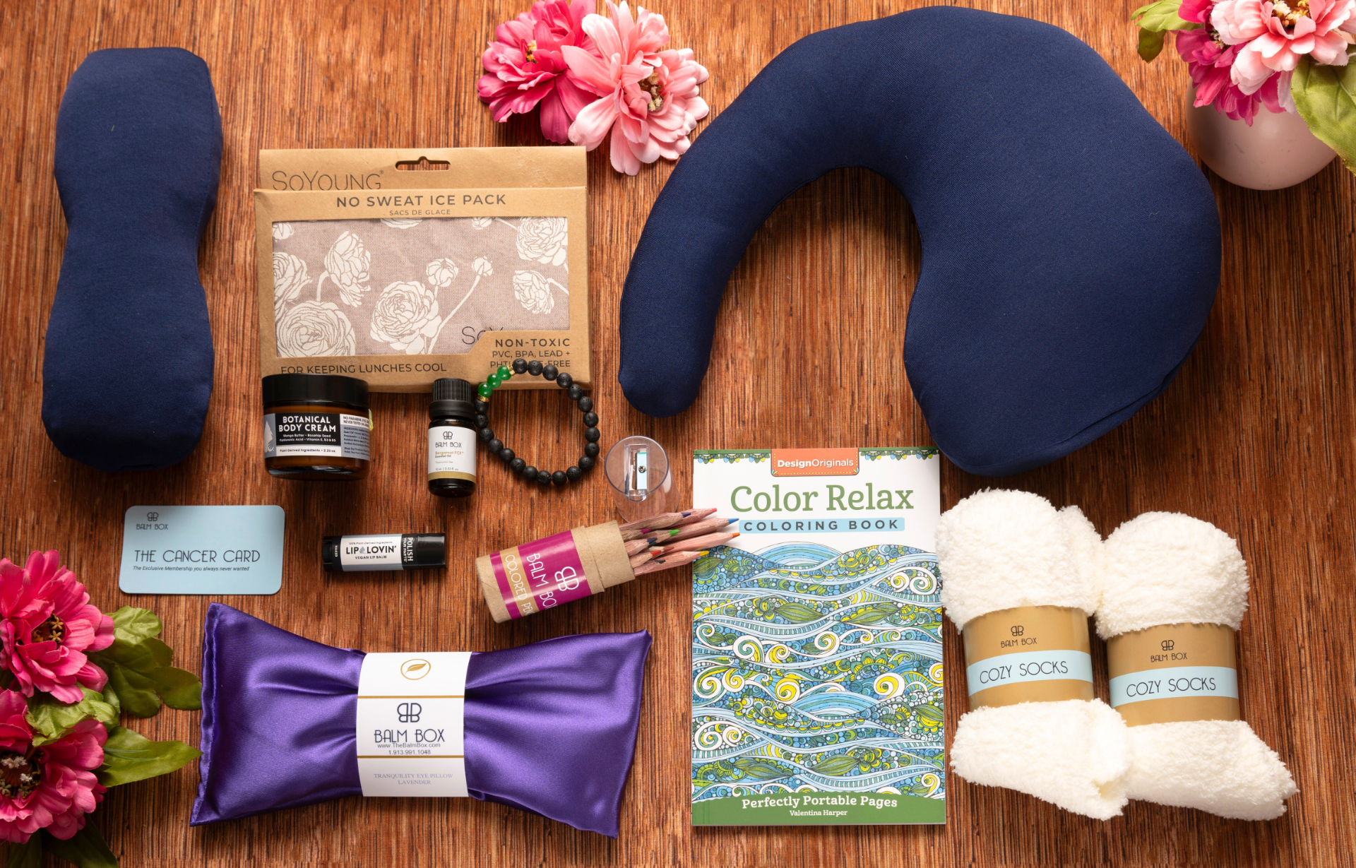 The Best Mastectomy Care Package features a blue U-shaped neck pillow, blue eye pillow, cozy white socks, coloring book with pencils, natural skincare products, and pink flowers—ideal comfort gifts beautifully arranged on a wooden surface.