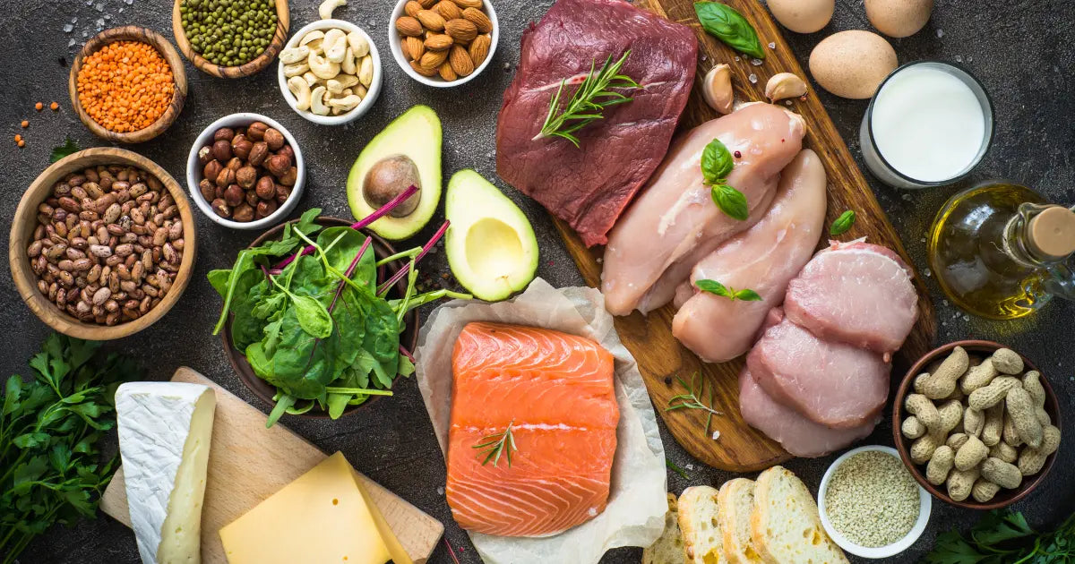 A variety of protein-rich foods, including raw salmon, chicken, red meat, eggs, cheese, assorted nuts, beans, spinach, avocado, milk, olive oil, and bread, arranged on a dark surface.