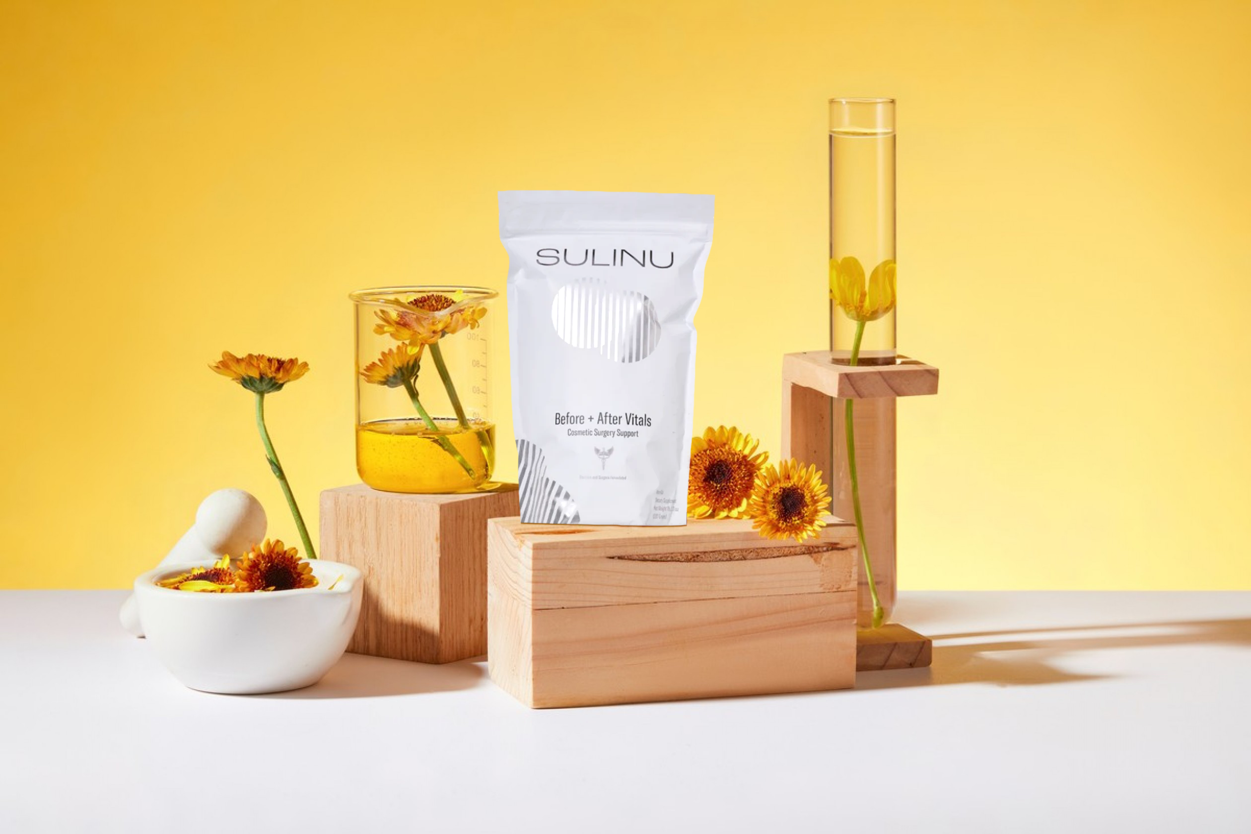 A white Sulinu supplement pouch stands on wooden blocks, surrounded by yellow flowers in glass containers and a bowl, all set against a bright yellow background.