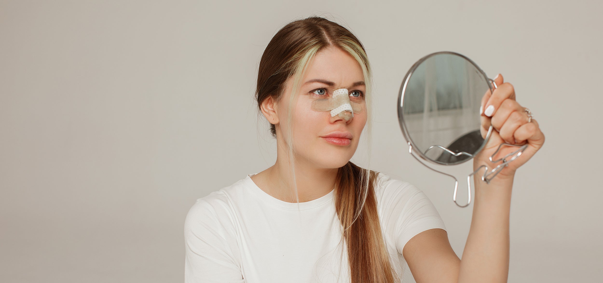 A woman looks at her nose bandage in the mirror. If you're preparing for a rhinoplasty, understanding the rhinoplasty swelling stages can be critical to keeping calm in your recovery.