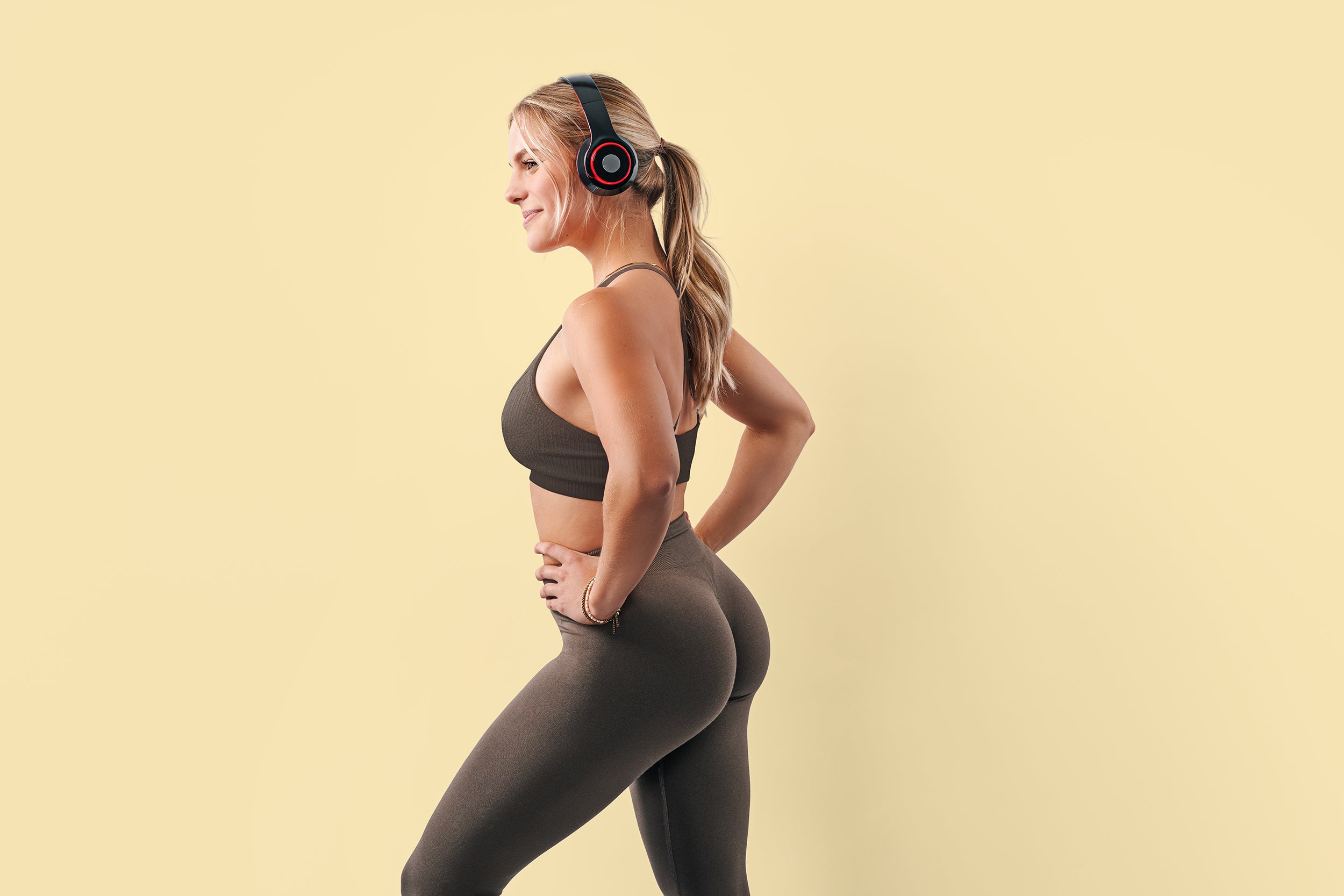 A woman in athletic wear and headphones stands with hands on hips against a light yellow background, looking to the side and smiling.