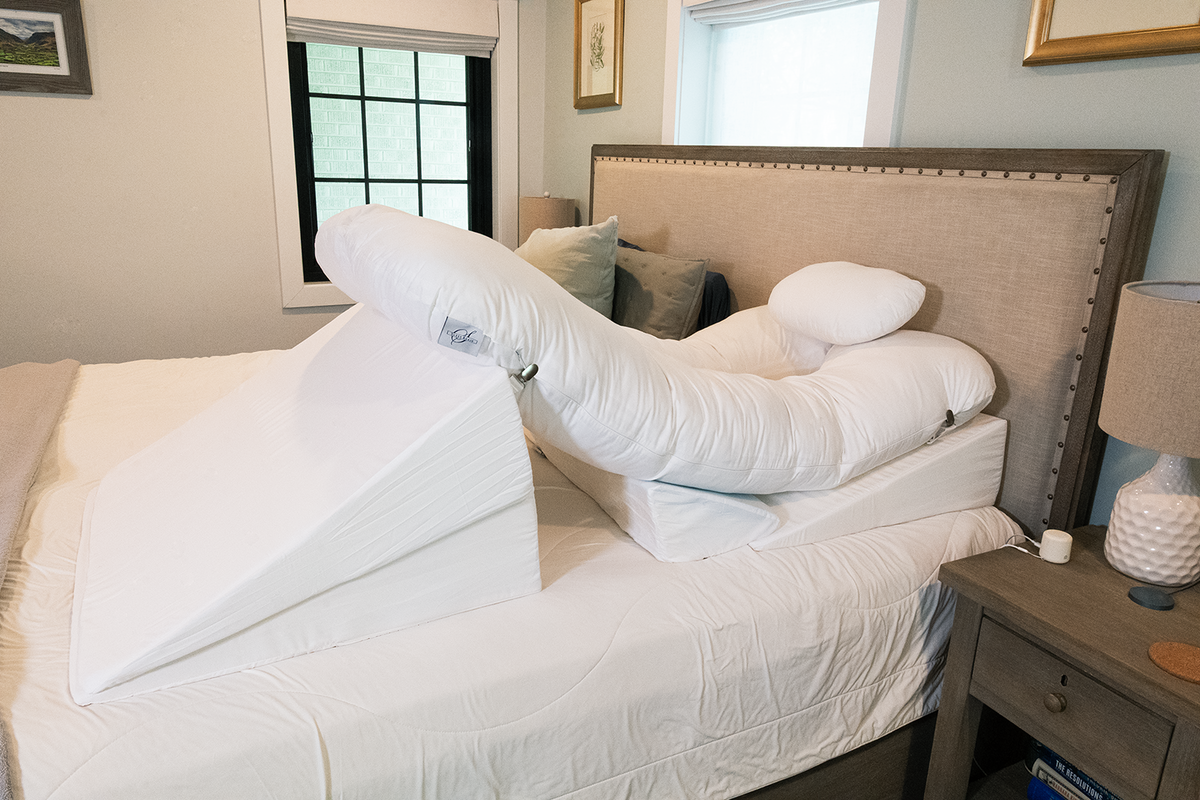 The BBL Add On Set, featuring a large white wedge pillow and supportive cushion, is arranged on a bed beside a beige headboard and nightstand with a lamp and pill bottle, creating a specialized pillow system to help someone sit up comfortably during their BBL recovery.