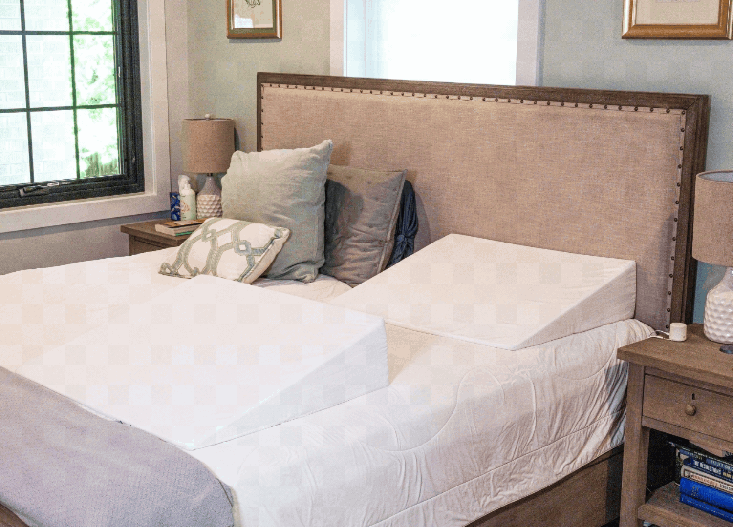A neatly made bed with a beige headboard features two white foam wedge pillows—one on each side—from the BBL Add On Set, perfect for BBL recovery after a Brazilian Butt Lift. Matching wooden nightstands with lamps and books sit on either side of the bed.