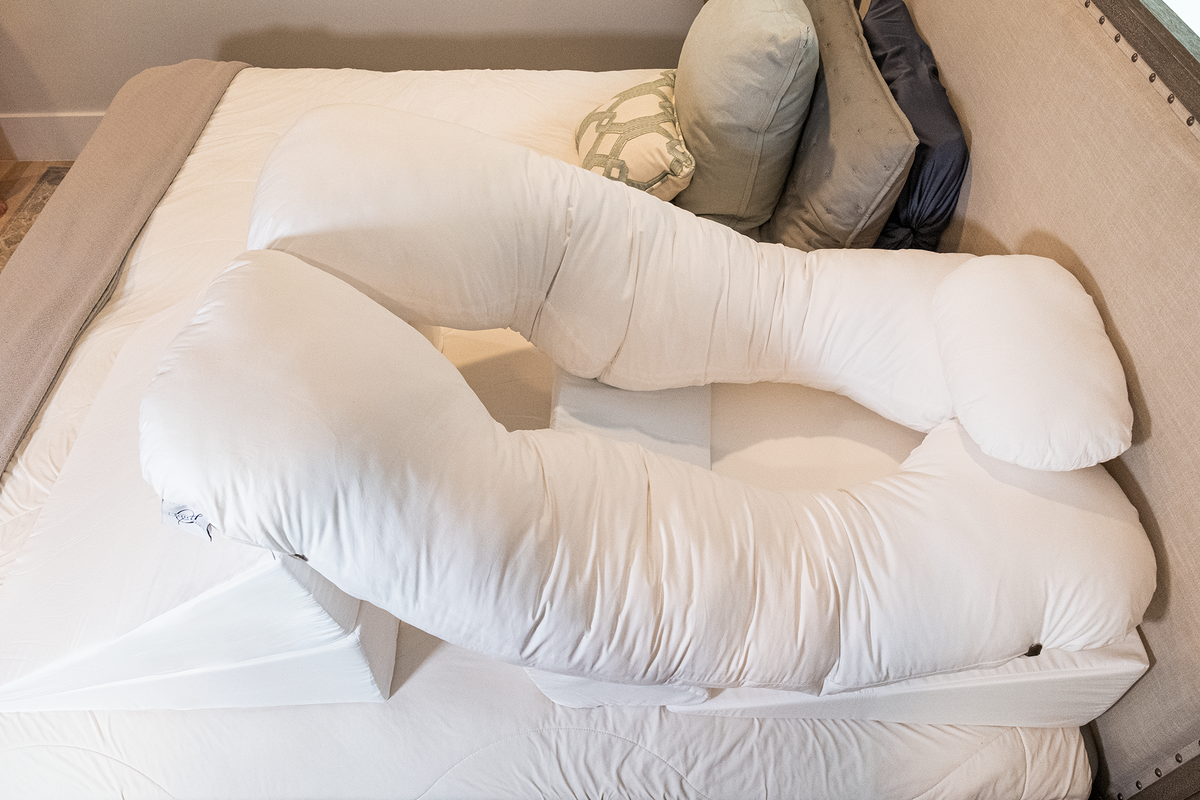 A BBL Add On Set—a large, white, U-shaped body pillow perfect for BBL recovery—rests on a bed with white sheets and several assorted pillows near the headboard.