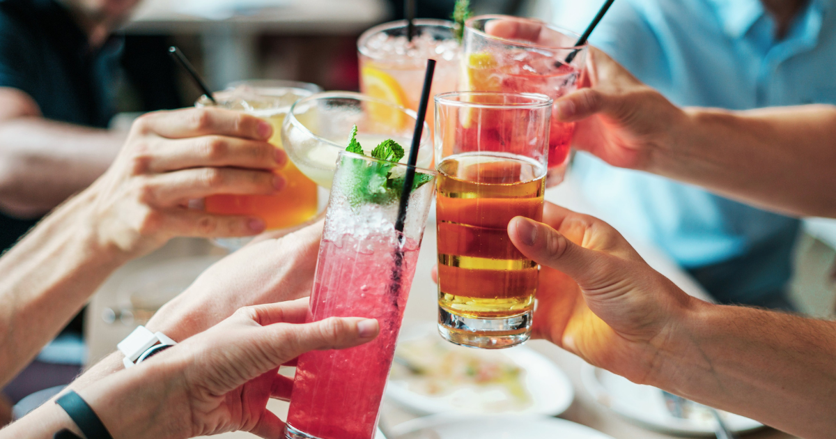 A group of people clinking various colorful drinks in glasses together for a toast, with hands raised around a table. The glasses contain cocktails and beer with ice and straws.