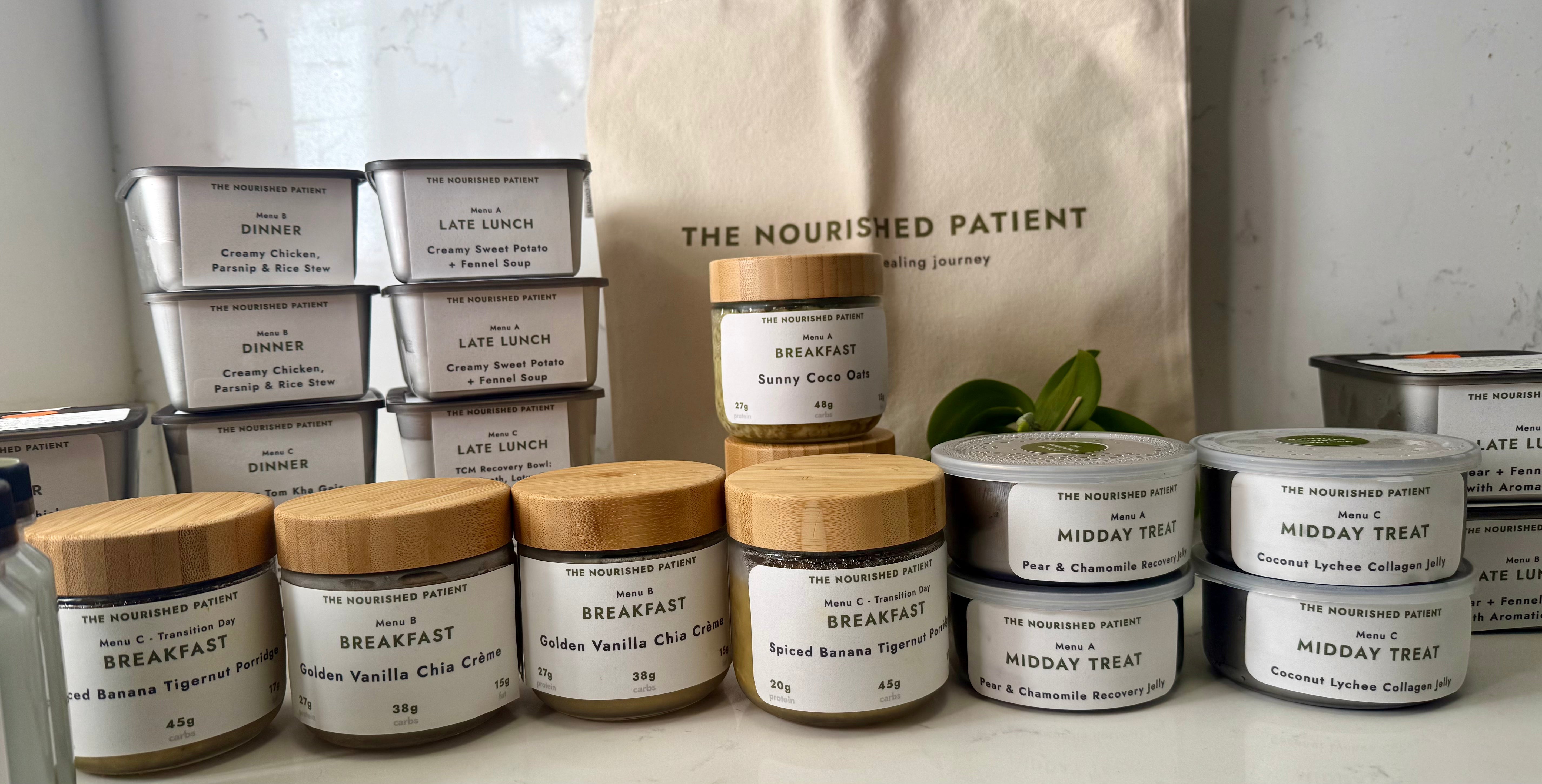 A display of labeled food containers from The Nourished Patient’s 7-Day Fat Transfer & Liposuction Recovery Meal Plan, arranged on a white surface with a branded beige tote bag and a small green plant in the background.