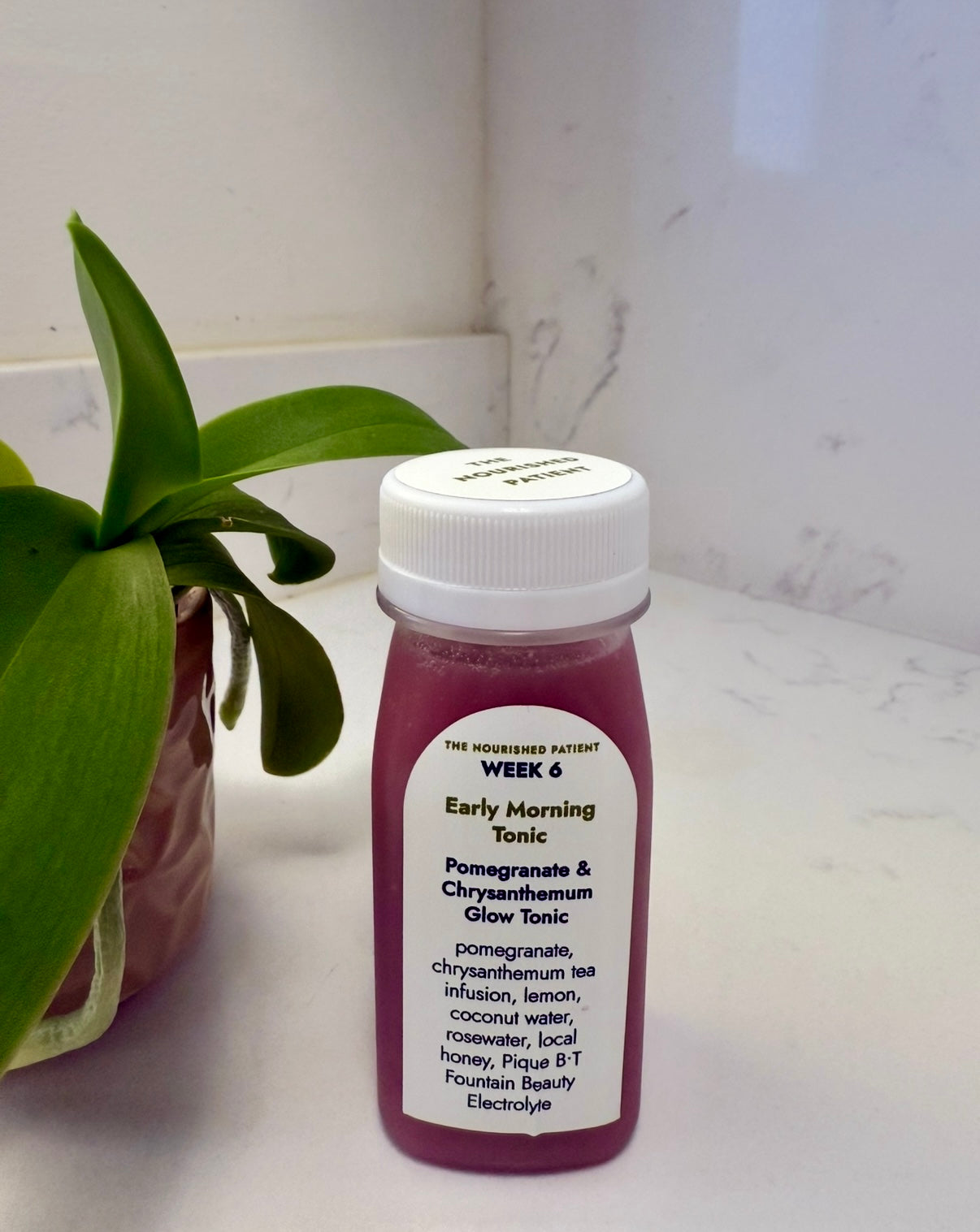 A small plastic bottle labeled Early Morning Tonic, containing pink liquid, sits on a white countertop near a green plant. The label highlights pomegranate and chrysanthemum tea, part of the 7-Day Facelift Recovery Meal Program to help reduce swelling.