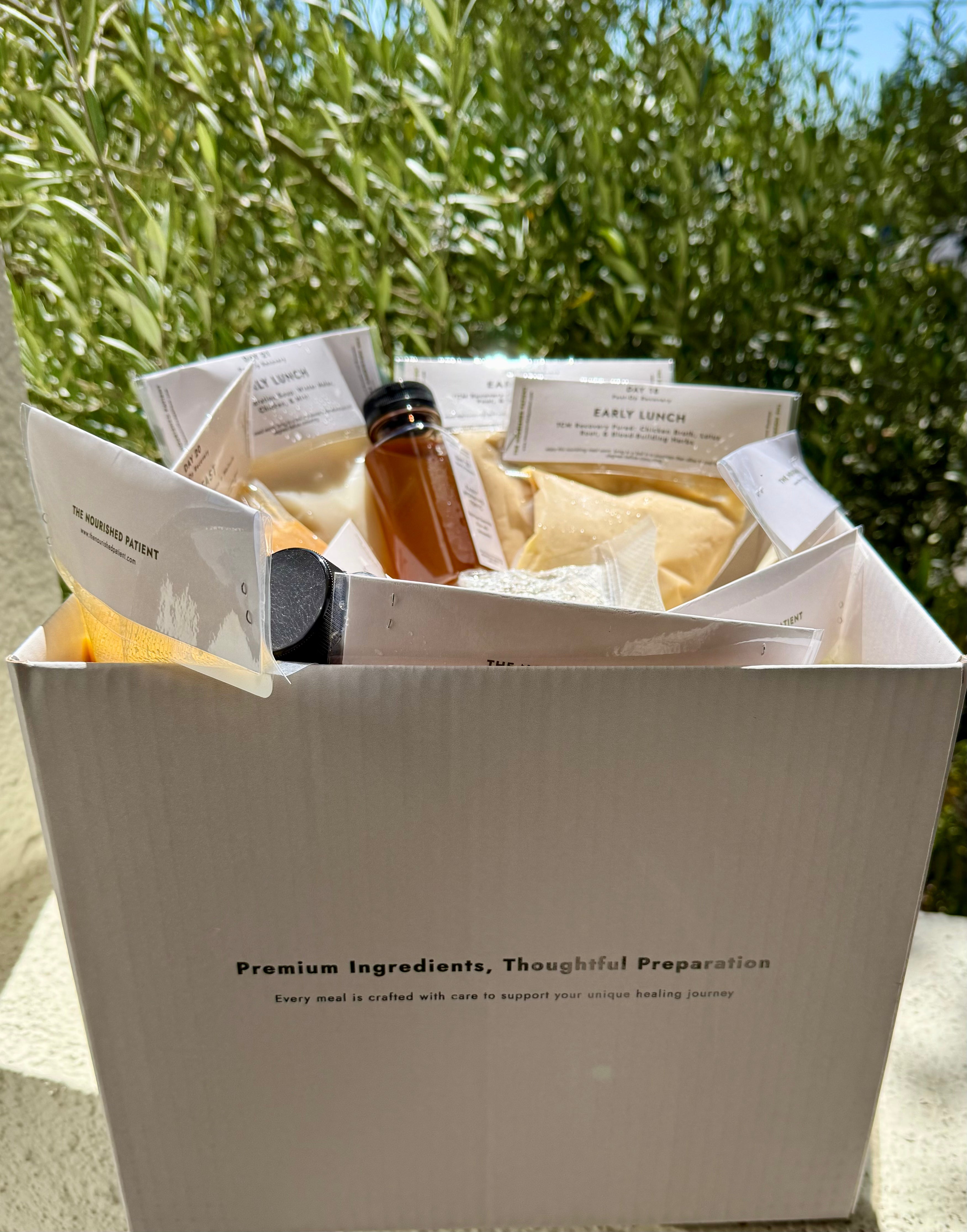 A white box labeled "7-Day Facelift Recovery Meal Program" holds packaged meals and drinks. Perfect for post-op delivery, it reads “Premium Ingredients, Thoughtful Preparation” and sits outside in sunlight with green foliage behind it.