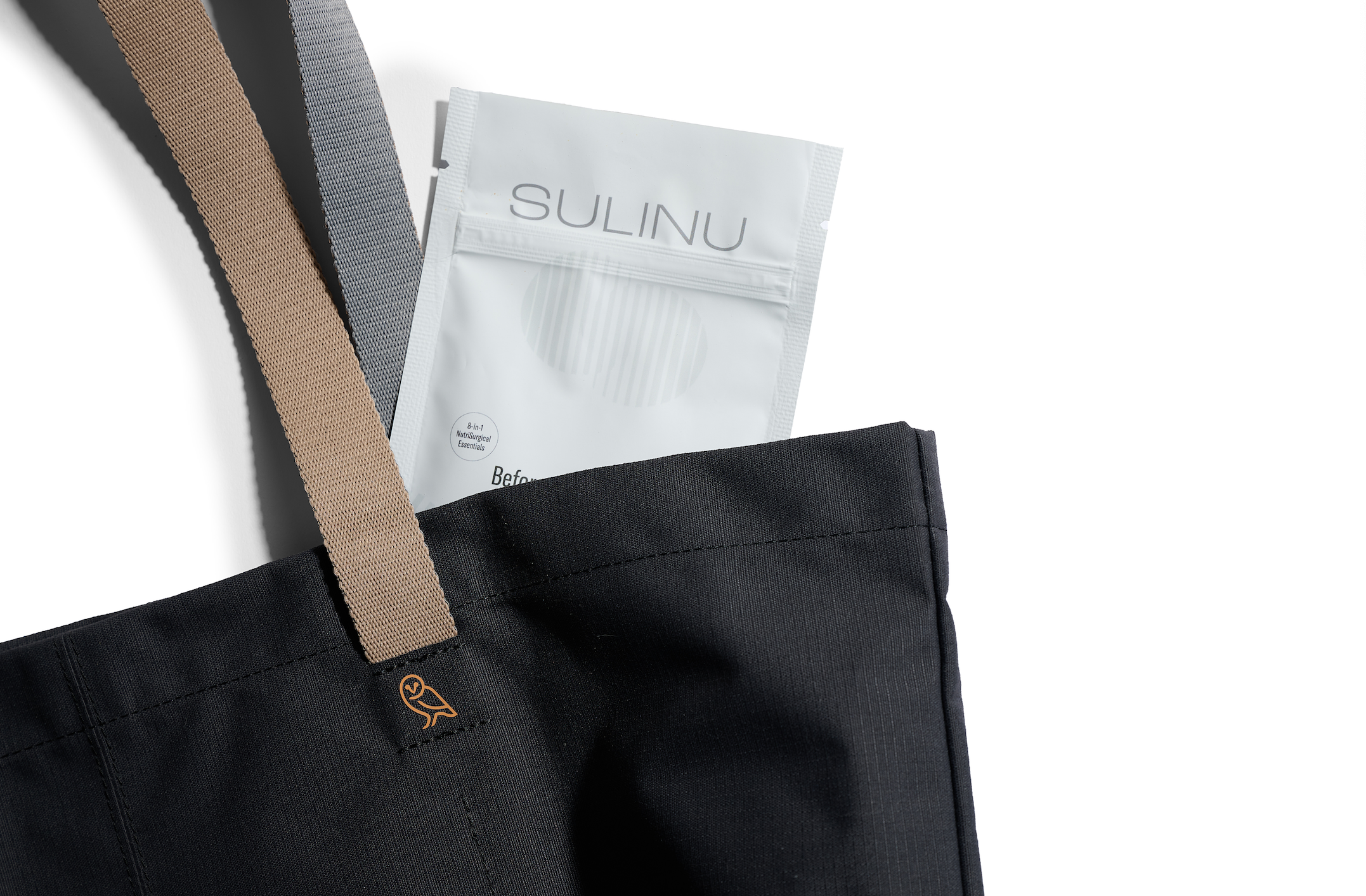 A black tote bag with beige and gray handles, featuring an embroidered owl logo, holds a white "SULINU" packet—Travel Packs- Set of 5- Single Serving Before + After Vitals—ideal for post-surgery vitamin needs.