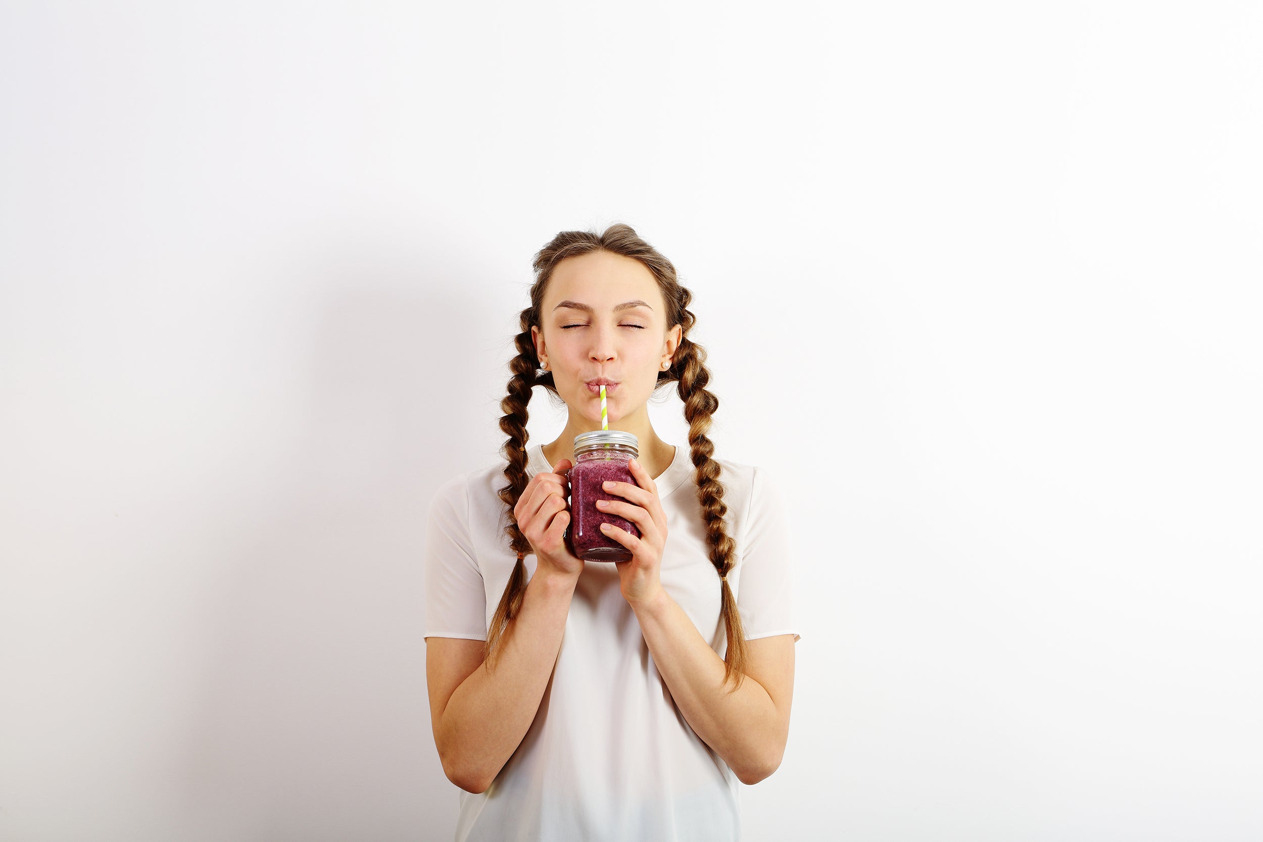 A young woman with braided hair, wearing a white T-shirt, is smiling with her eyes closed while drinking a purple smoothie from a glass jar using a straw, standing against a plain white background.