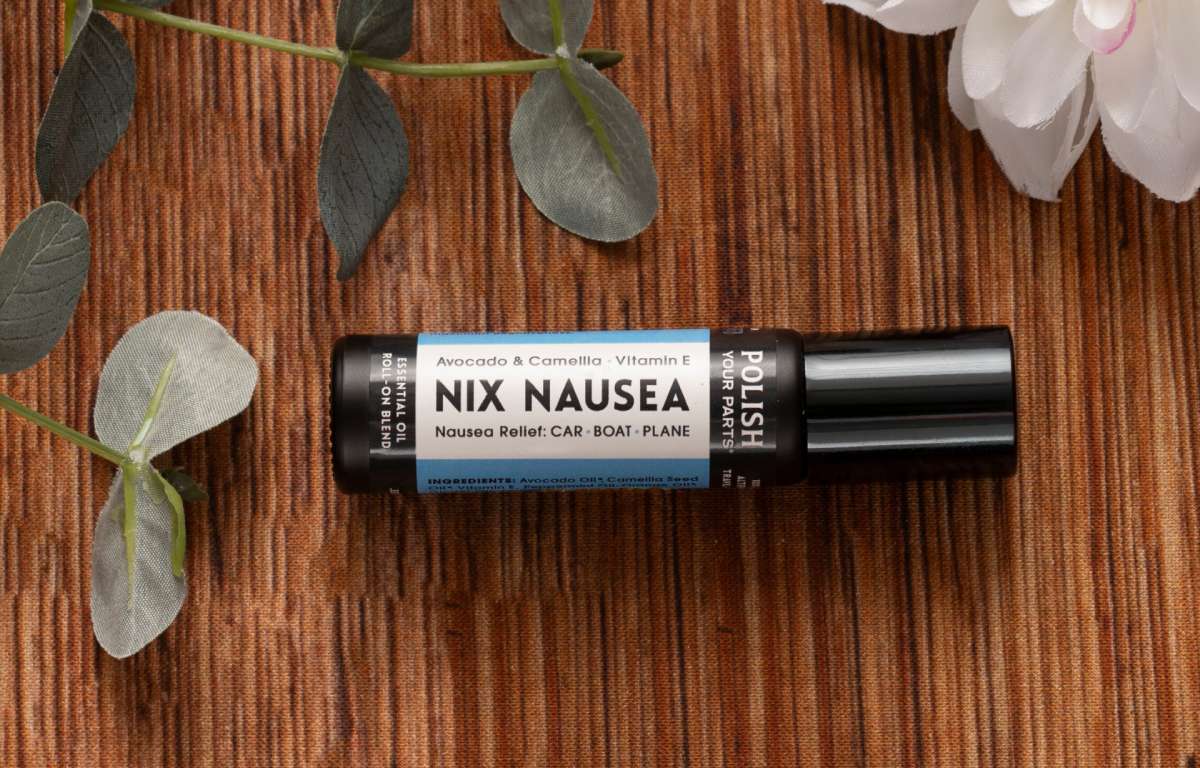 A bottle from The Perfect Little Anti-Nausea Box rests on a brown textured surface, surrounded by green eucalyptus leaves and a white flower. Its label highlights avocado, camellia, and vitamin E—perfect for a cancer patient gift box.