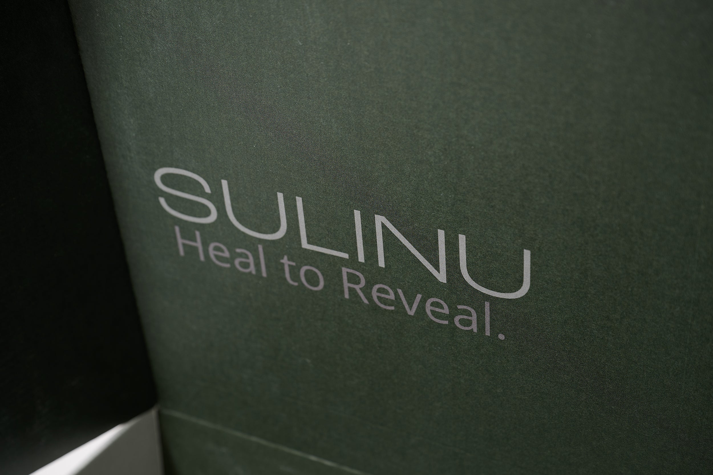 Close-up of a dark green box with "SULINU Heal to Reveal." in large, light text, featuring The VIP Surgical Recovery Kit to support your cosmetic surgery recovery journey.